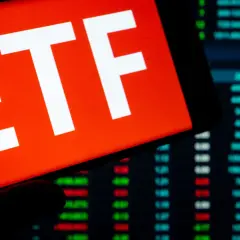 Ein Smartphone zeigt "ETF" auf rotem Grund. Im Hintergrund sind Börsenkurse zu sehen. Ein ETF steuerfrei nach 10 Jahren ist ein Traumziel für Anleger.
