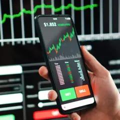 Ein Smartphone zeigt einen Aktien-Tracker mit einem steigenden Chart. Daneben sind Kauf- und Verkaufsbuttons zu sehen.