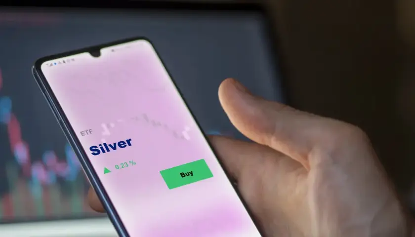 Hand hält Smartphone mit ETF Silber Kursanzeige. Ein grüner "Buy"-Button ist sichtbar, daneben ein Pfeil nach oben und 0,23 %.