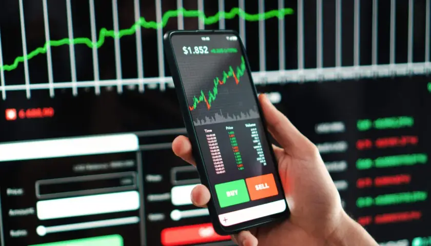 Ein Smartphone zeigt einen Aktien-Tracker mit einem steigenden Chart. Daneben sind Kauf- und Verkaufsbuttons zu sehen.