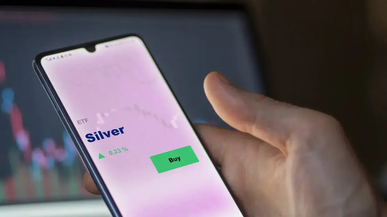 Hand hält Smartphone mit ETF Silber Kursanzeige. Ein grüner "Buy"-Button ist sichtbar, daneben ein Pfeil nach oben und 0,23 %.