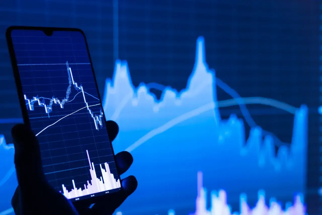 Ein Handy zeigt einen Börsenchart, während im Hintergrund ein größerer Chart zu sehen ist. Ein STP Broker ermöglicht den direkten Marktzugang.