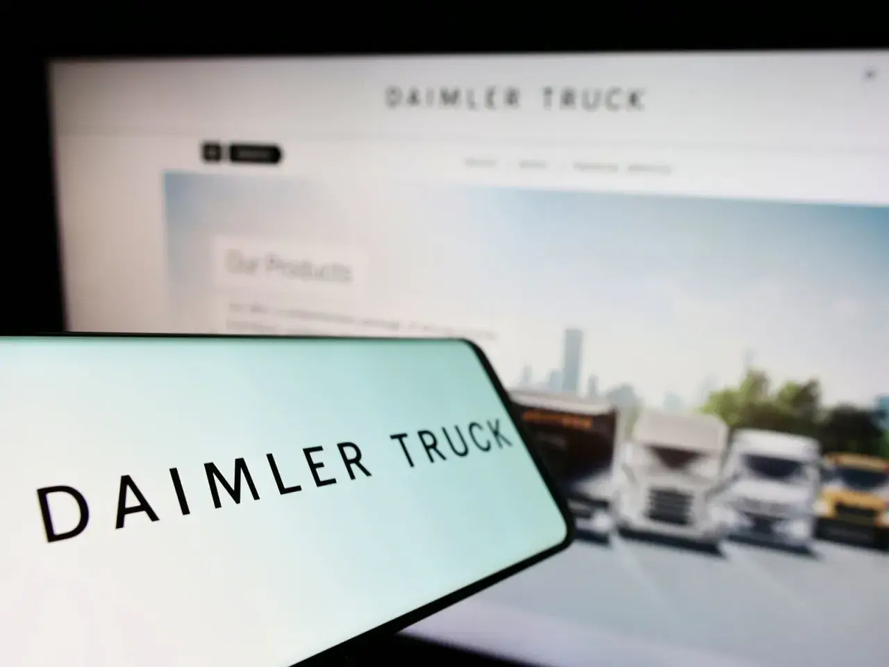 Daimler Truck Logo auf einem Smartphone, im Hintergrund ein Bildschirm mit LKW-Bildern und der Aufschrift "Our Products". Die Daimler Truck Aktie Kurs Prognose im Fokus.